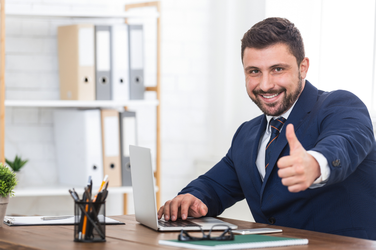 A man in a suit, thumbs up with a laptop in front of him