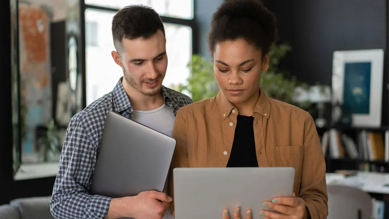 A woman and a man holding laptops, standing side by side, looking down at one of the screens together, focused and engaged.