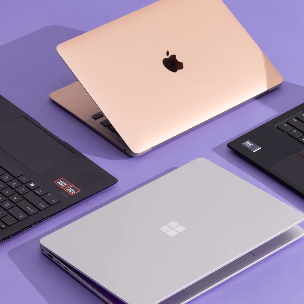 A flat-lay arrangement of four modern laptops on a purple background, including a rose gold MacBook, a silver Microsoft Surface laptop, and two black Windows laptops.